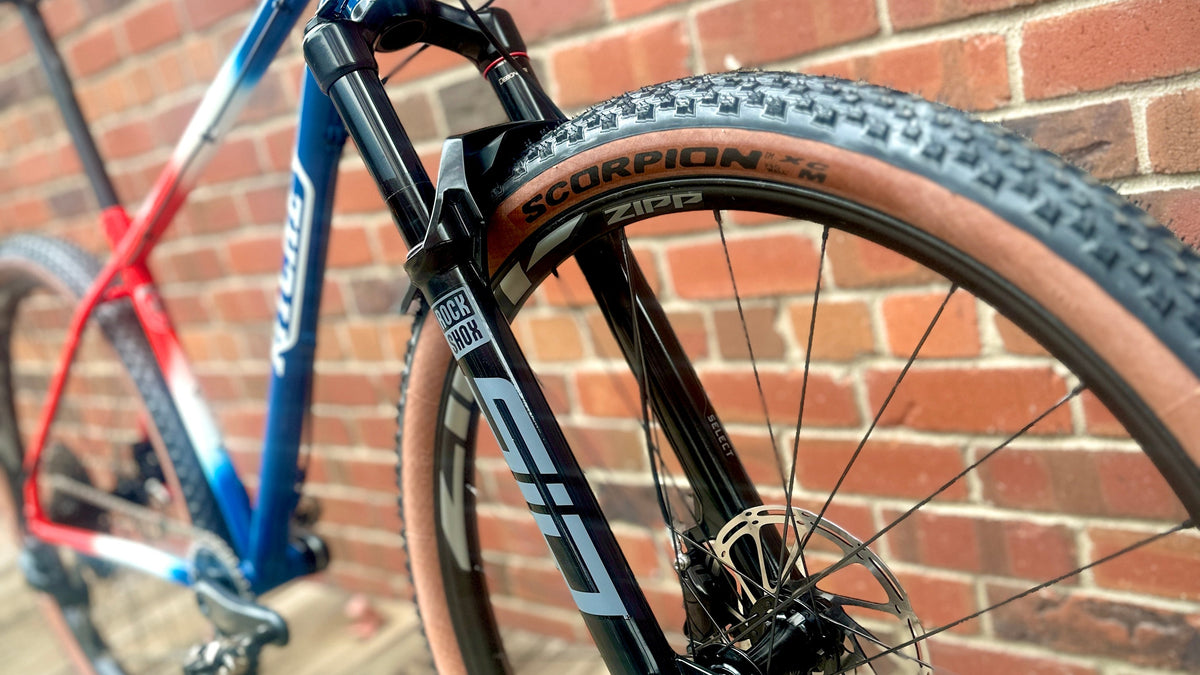 Close-up of a mountain bike's front wheel and suspension against a red brick wall and wooden floor. The bike frame is blue, white, and red, with a black front fork and knobby tire.
