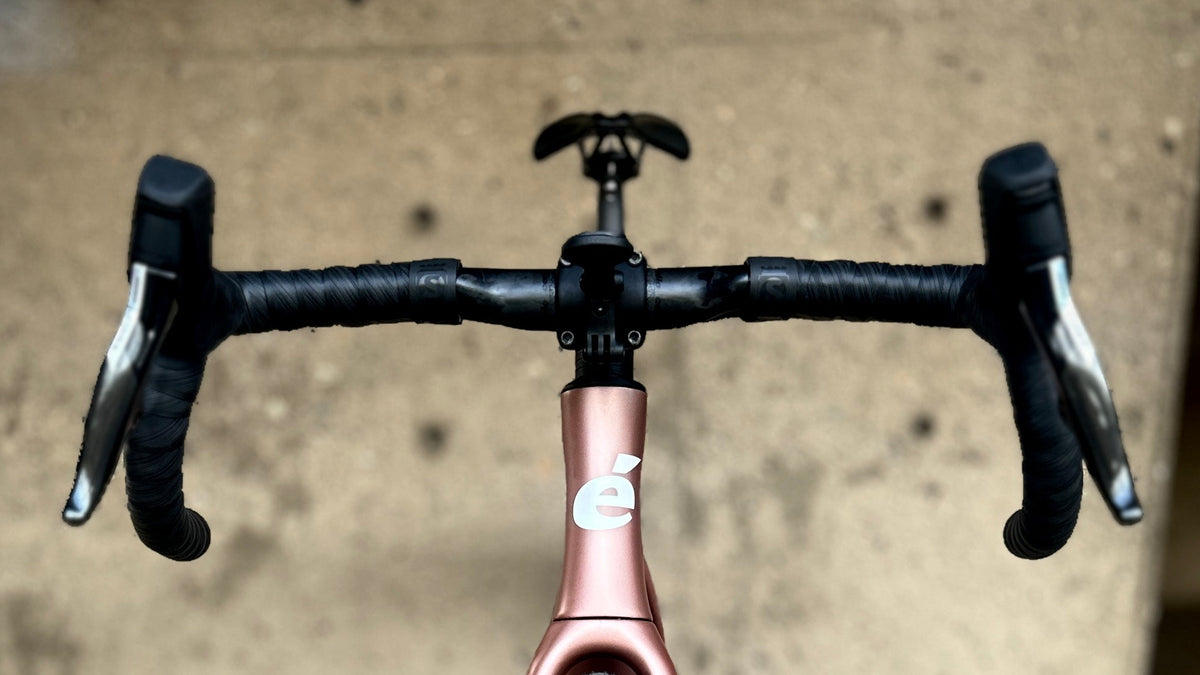 A drop-bar bicycle photographed head-on from the front, with a metallic rose-pink frame, black handlebars, black tires, and a black saddle, set on a weathered wooden deck in front of a concrete wall.