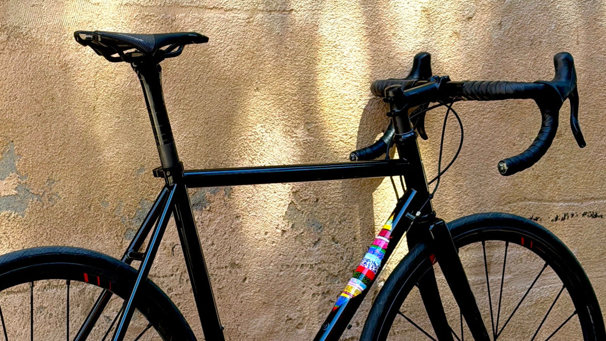 A black road bicycle with drop handlebars and thin tires leans against a textured beige wall under dappled sunlight and shadows.