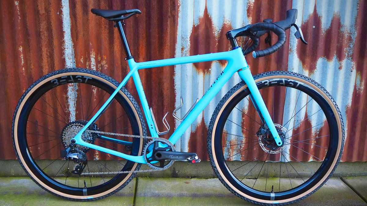 A light blue gravel bicycle with black components and tan sidewall tires, parked against a corrugated metal wall.