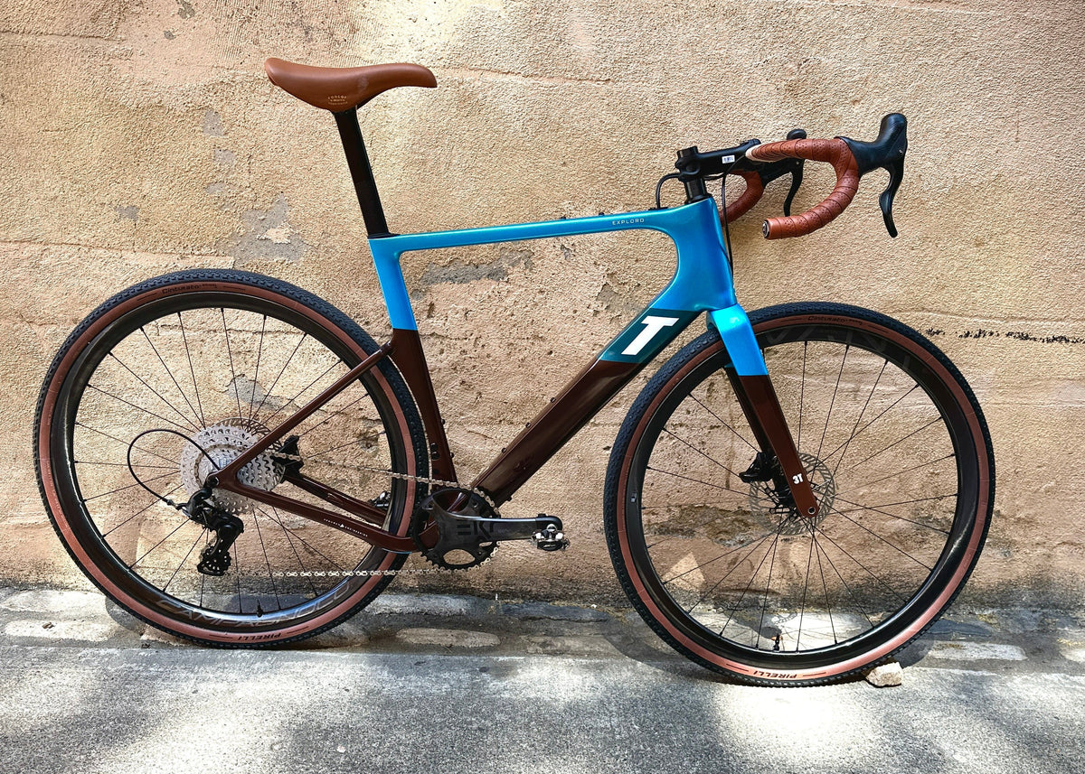 A modern drop-bar road bicycle with a bright blue frame and dark brown carbon fiber sections, parked against a textured beige concrete wall.